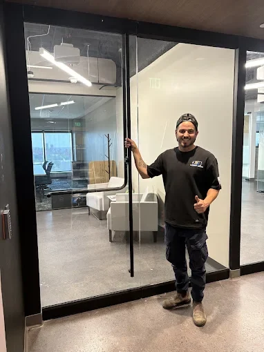 JDM technician Yuval standing beside a full-height glass office door he has just serviced, holding the vertical pull handle in a bright commercial lobby