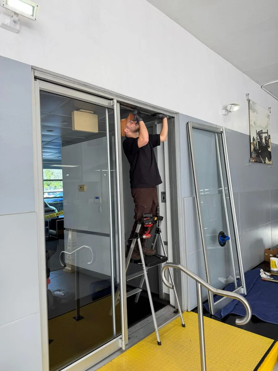 JDM technician Yuval on a step ladder working at the head of an aluminum commercial storefront door frame, with a removed door panel resting against the wall behind him