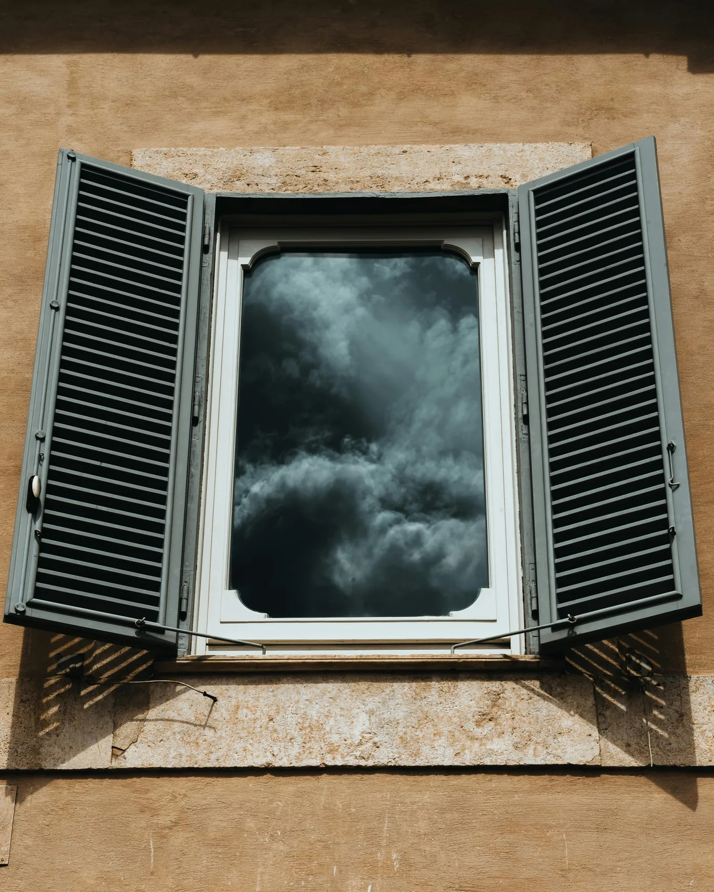 Window with louvered shutters open against a tan stucco wall and stormy sky, the alternative many Florida homeowners weigh against impact glass